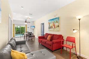 Ein Wohnzimmer mit einem grauen Ecksofa, einem roten Sofa und einem roten Sessel. An der Wand hängt ein Gemälde, es gibt einen Deckenventilator und eine Stehlampe.