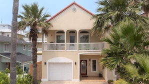A two-story house with a front garage, a balcony, and palm trees. - Dog-Friendly Townhome • Pool & Hot Tub • 5 Minute Walk to Beach (South Padre Island)