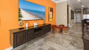 A living room with a large TV, a wooden entertainment unit, a dining area with chairs, and a kitchen in the background. - Spacious Townhouse • Private Pool • 2 Minute Walk to the Beach (South Padre Island)