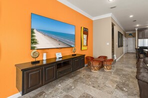 A living room with a large TV, a wooden entertainment unit, a dining area with chairs, and a kitchen in the background. - Spacious Home w/ Pool, 1/2 Block to Beach (South Padre Island)
