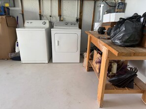 Ein Waschbereich mit zwei weißen Waschmaschinen, einem Holztisch mit einer schwarzen Müllsäcke und einem Durchlauferhitzer an der Wand.