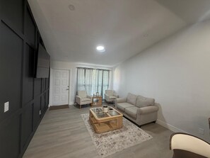 Ein Wohnzimmer mit einer Couch, zwei Sesseln, einem Couchtisch und einem Fernseher an der Wand.