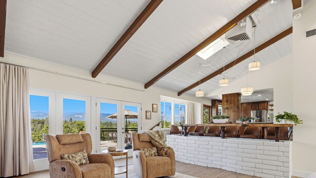 A spacious living area with a high ceiling, large windows, and a view of mountains. There are two leather armchairs, a wooden coffee table, and a kitchen area with wooden cabinets and a bar. - New! Yosemite Starsong with pool/hot tub/EV/huge views (Coarsegold)