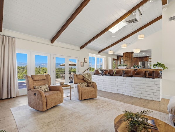 A spacious living area with a high ceiling, large windows, and a view of mountains. There are two leather armchairs, a wooden coffee table, and a kitchen area with wooden cabinets and a bar.
