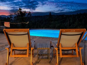 Two wooden lounge chairs with mesh backs facing a swimming pool, surrounded by a deck with a warning sign.