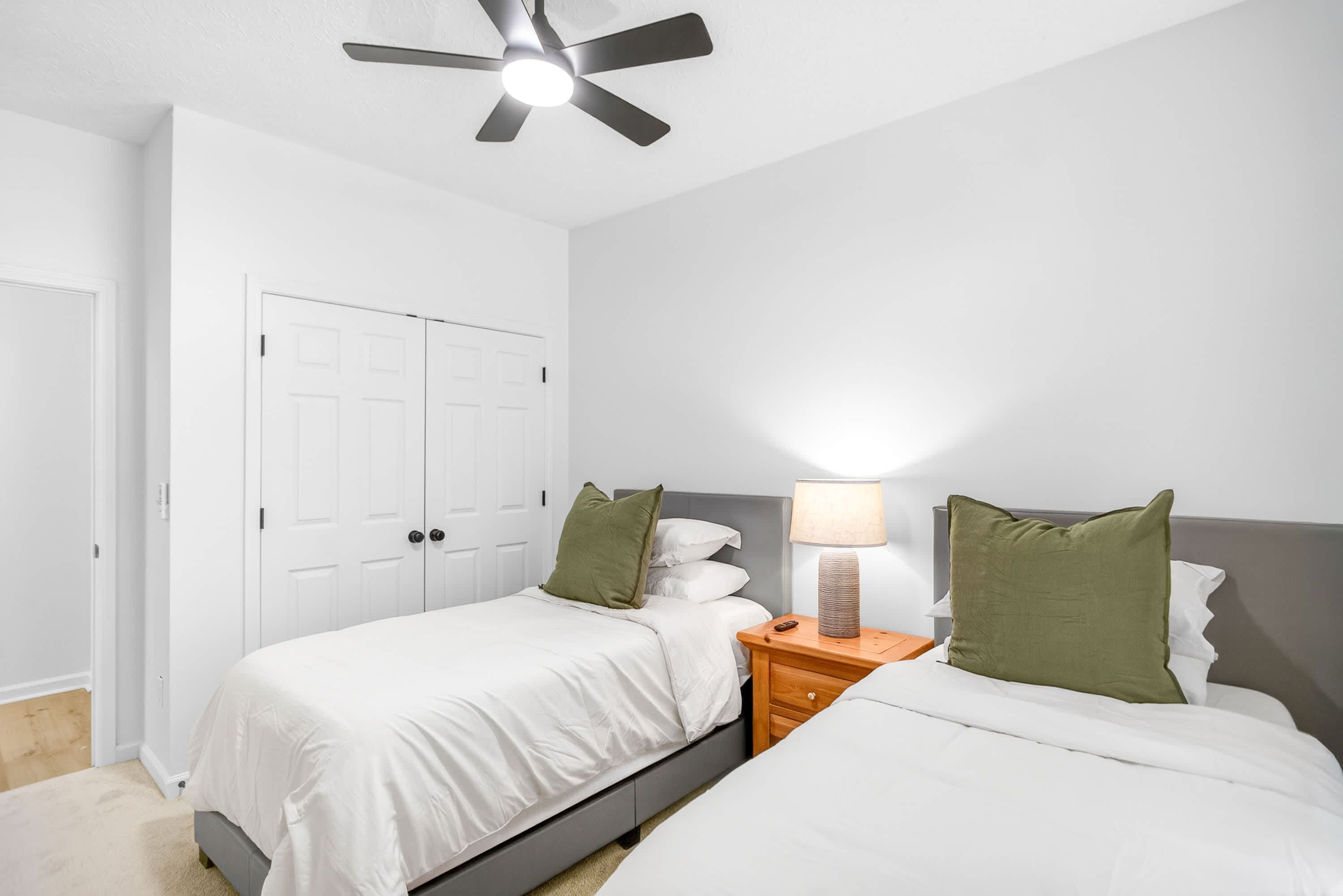 A bedroom with two beds, a ceiling fan, a bedside table with a lamp, and a wardrobe.