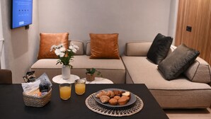 A living room with a sofa, a coffee table with breakfast items, and a television on the wall. - Cocoon Athenian Riviera by Neuvel (Vari-Voula-Vouliagmeni)