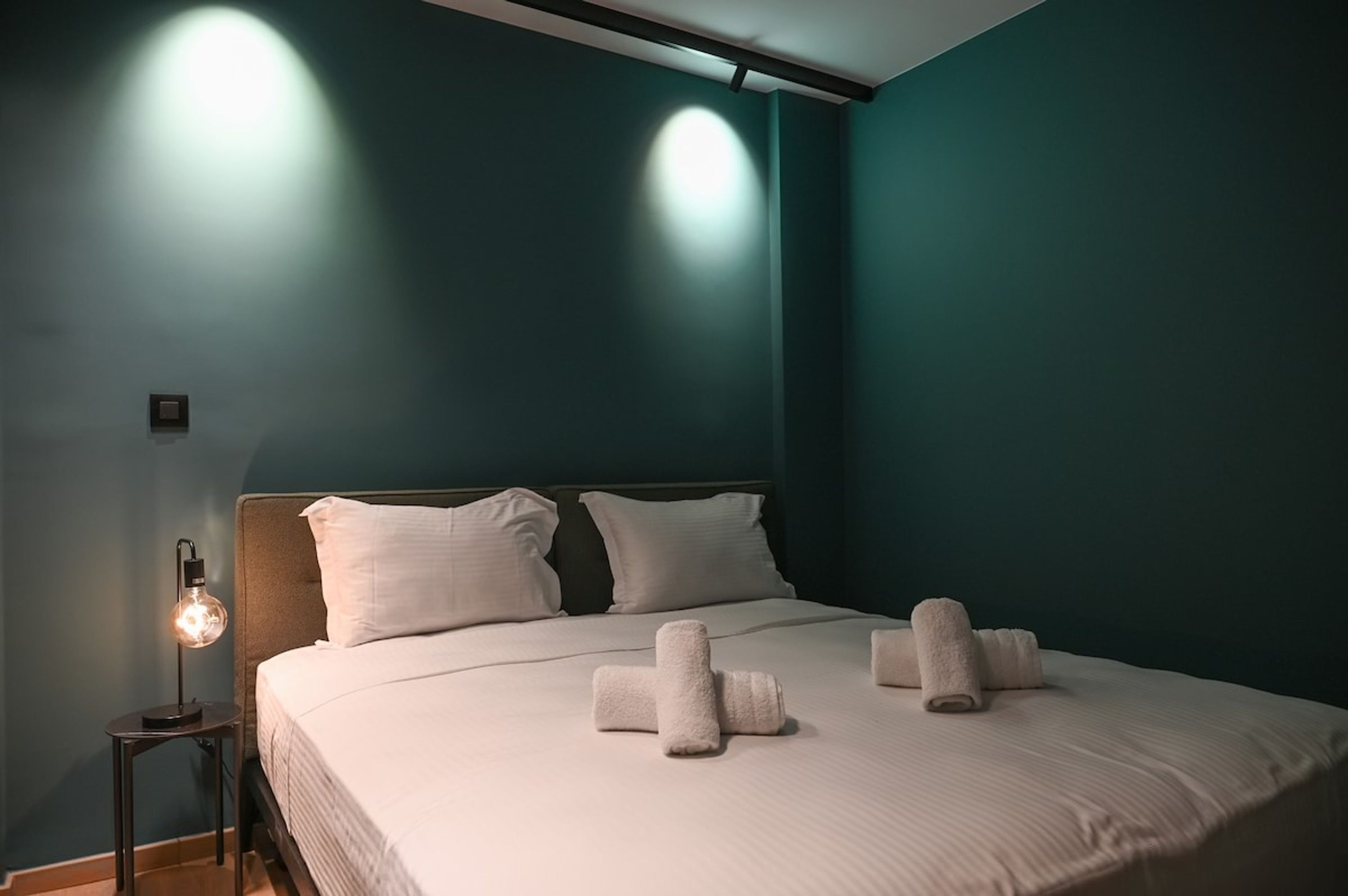 A neatly made bed with white linens and towels, a bedside lamp, and a wall-mounted light fixture.