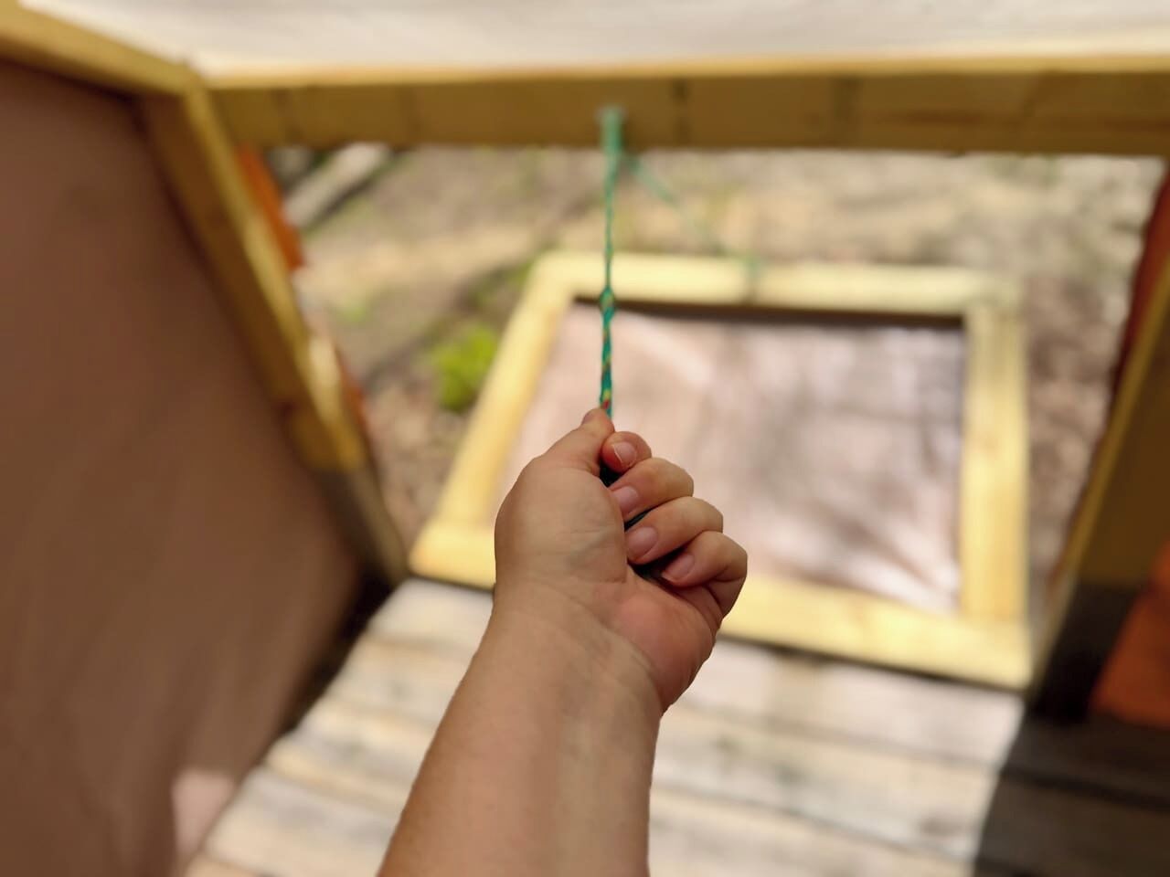 A hand holding a green string, with a wooden structure and a blurred background.