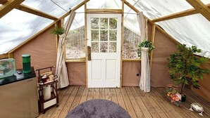 Une tente confortable avec une structure en bois, une porte blanche et une petite table sur laquelle se trouvent un ventilateur et une bouilloire.
