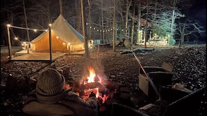Un espace aménagé avec des tentes et un feu de camp, entouré d’arbres et illuminé de guirlandes lumineuses.