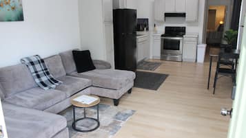 Una sala moderna con un sofá seccional gris, una mesita redonda y una zona de cocina con armarios blancos y electrodomésticos de acero inoxidable.