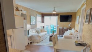 Een moderne woonkamer met een bank, een eettafel met stoelen, een aan de muur bevestigde televisie en een keuken met een koelkast.