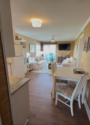 A modern living room with a sofa, a dining table with chairs, a TV mounted on the wall, and a kitchen area with a refrigerator. - Beach Haven •  Ground Floor • Walk to Beach • Pool + Fishing Dock • Gated (Murrells Inlet)