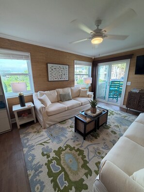 A living room with a beige sofa, a coffee table with a plant, a ceiling fan, and a large window with a view. - Beach Haven •  Ground Floor • Walk to Beach • Pool + Fishing Dock • Gated (Murrells Inlet)