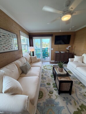 A living room with a beige sofa, a coffee table, a TV, and a ceiling fan. - Beach Haven •  Ground Floor • Walk to Beach • Pool + Fishing Dock • Gated (Murrells Inlet)
