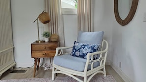 Une chaise en osier blanche avec un coussin à motifs floraux, une table d’appoint en bois avec une lampe, et un mur blanc orné d’un miroir et de prises électriques.