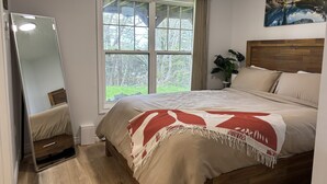 A bedroom with a bed, a window with curtains, a mirror, and a painting on the wall.