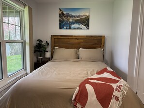 Ein Schlafzimmer mit einem hölzernen Kopfteil, einem Bett mit beige farbenem Bettzeug, einer roten und weißen Decke sowie einem Fenster mit Blick auf Grünflächen. An der Wand hängt ein Gemälde einer Berglandschaft.