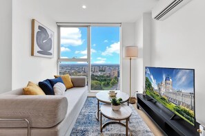 Un salon moderne avec un canapé, une table basse ronde, un téléviseur et une grande fenêtre donnant sur le paysage urbain.