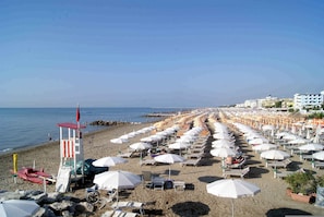 Ein Strand mit Reihen weißer Sonnenschirme und Liegestühle, einem Rettungsturm und Gebäuden im Hintergrund.