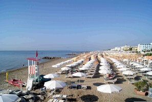 A beach with rows of white umbrellas and sun loungers, a lifeguard tower, and buildings in the background. - Holiday apartment mit Klimaanlage und Balkon (Bibione)