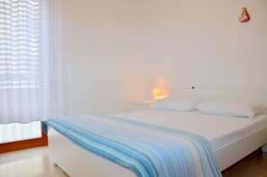 A bedroom with a bed, a nightstand, a lamp, and a window with curtains. - Holiday apartment mit Klimaanlage und Balkon (Bibione)