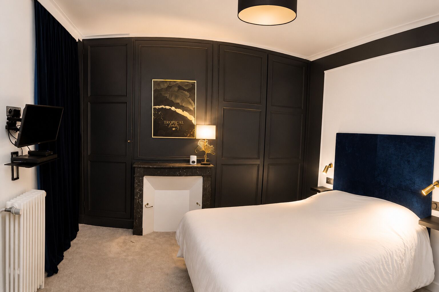 A hotel room with a bed, a desk, a chair, a radiator, a television, and a wall-mounted artwork.
