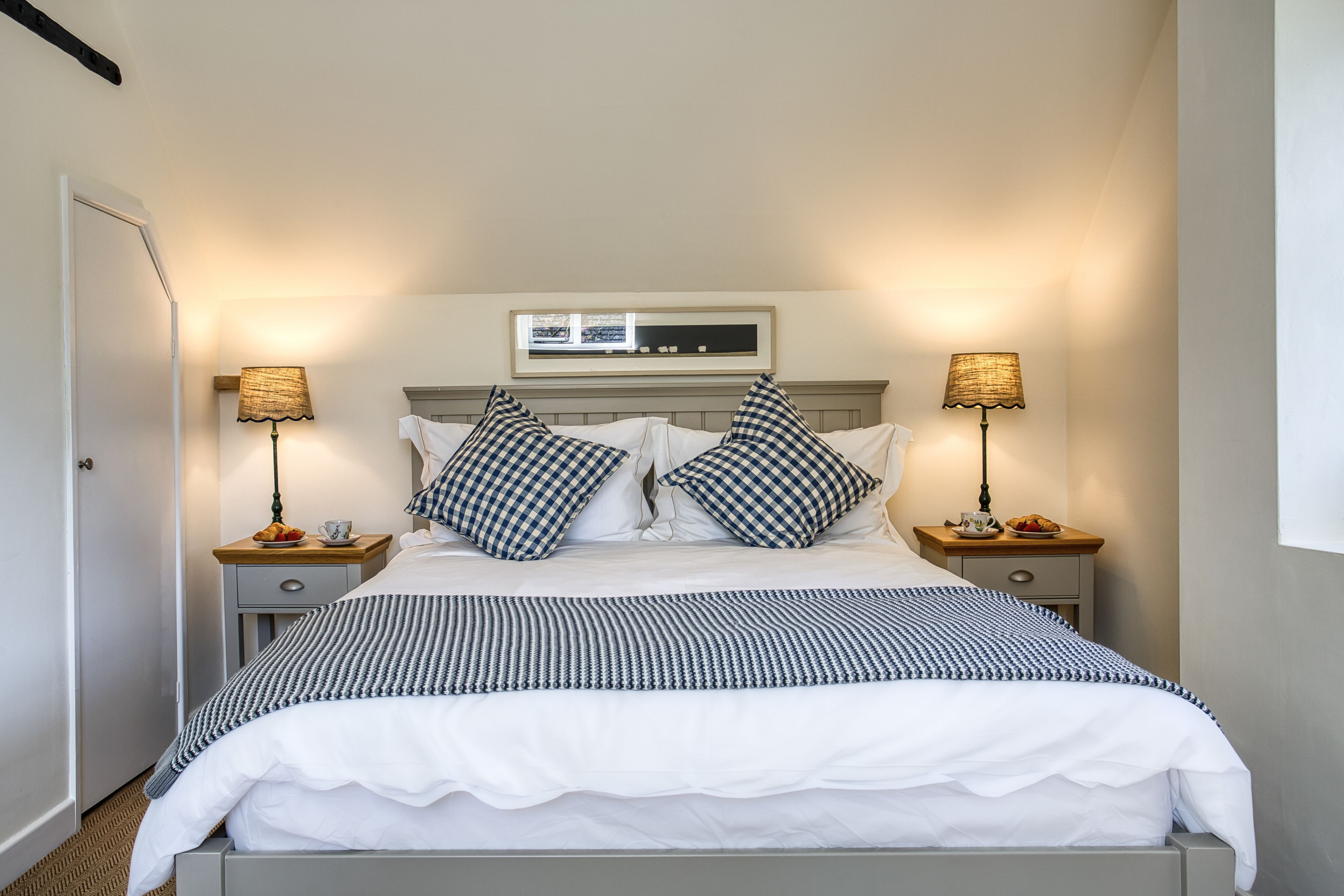 A neatly made bed with checkered pillows, two bedside tables with lamps, a framed picture above the bed, and a window with blinds.