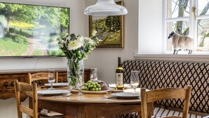 Un comedor con una mesa de madera dispuesta para cuatro personas, un jarrón con flores, una botella de vino y un televisor que muestra una vista panorámica.
