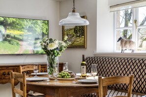 A dining area with a wooden table set for four, a vase with flowers, a bottle of wine, and a television displaying a scenic view. - Roundhill Farm Cottage sleeps 2 guests  in 1 bedroom (Naunton)