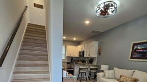 A modern living room with a staircase, a kitchen area, and a sofa with a beach-themed pillow.