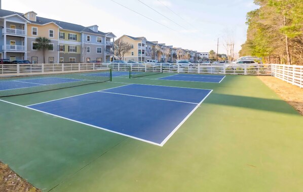 A tennis court with a net, surrounded by a white fence, and residential buildings in the background.