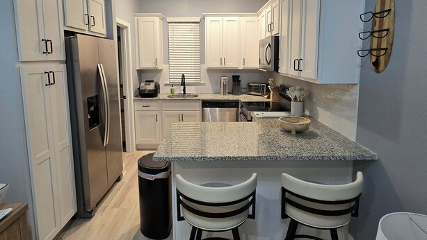A modern kitchen with white cabinets, a granite countertop, stainless steel appliances, and a bar area with two stools.