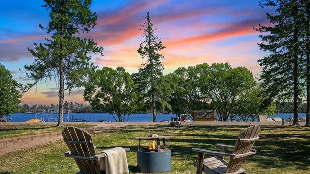 Dos sillas Adirondack, un brasero y una vista de un lago con árboles y un muelle al fondo.
