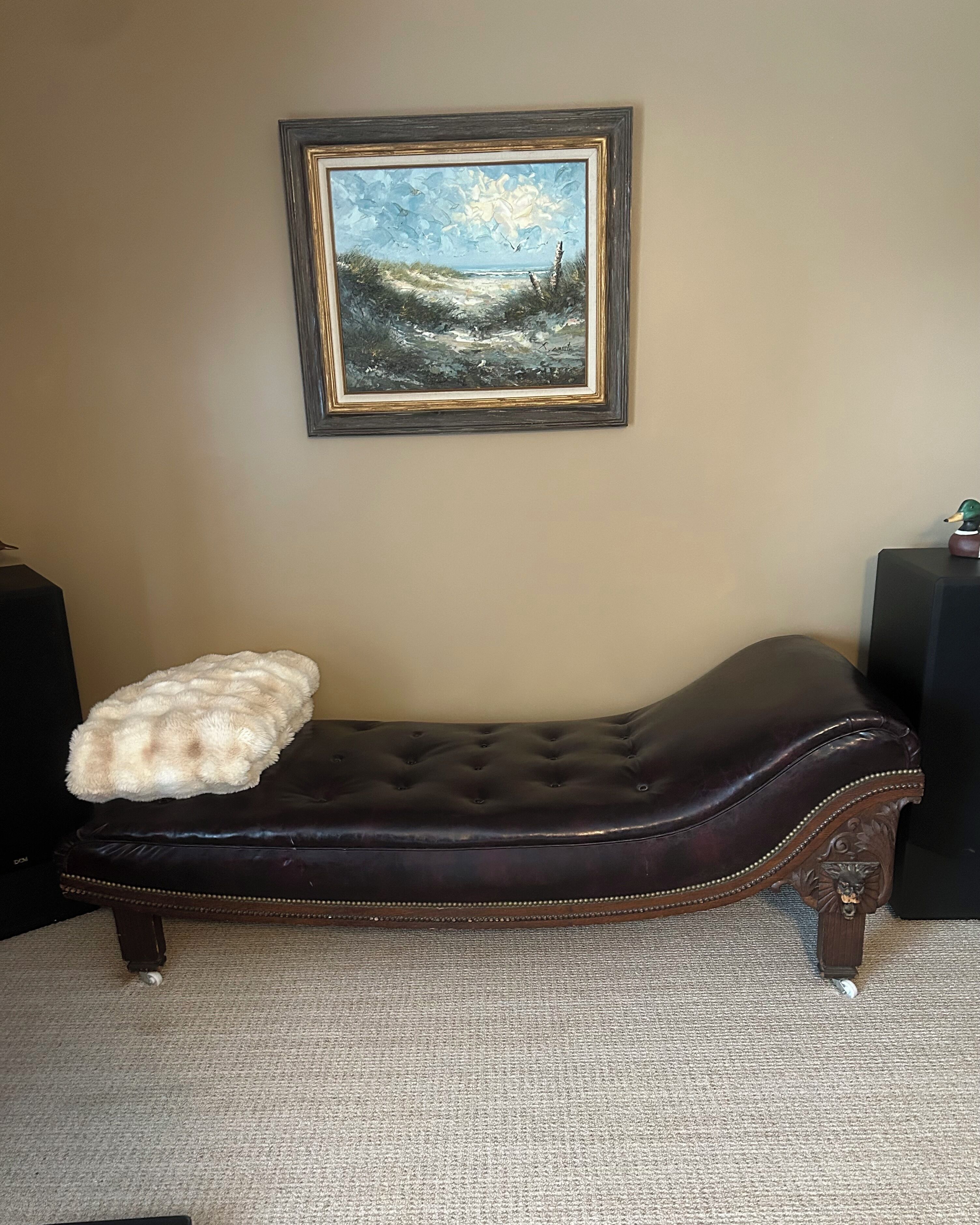 A dark brown upholstered bench with a tufted backrest and decorative trim, a white fluffy throw pillow, and a framed painting of a landscape on the wall.