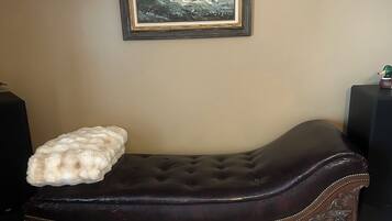 A dark brown upholstered bench with a tufted backrest and decorative trim, a white fluffy throw pillow, and a framed painting of a landscape on the wall.