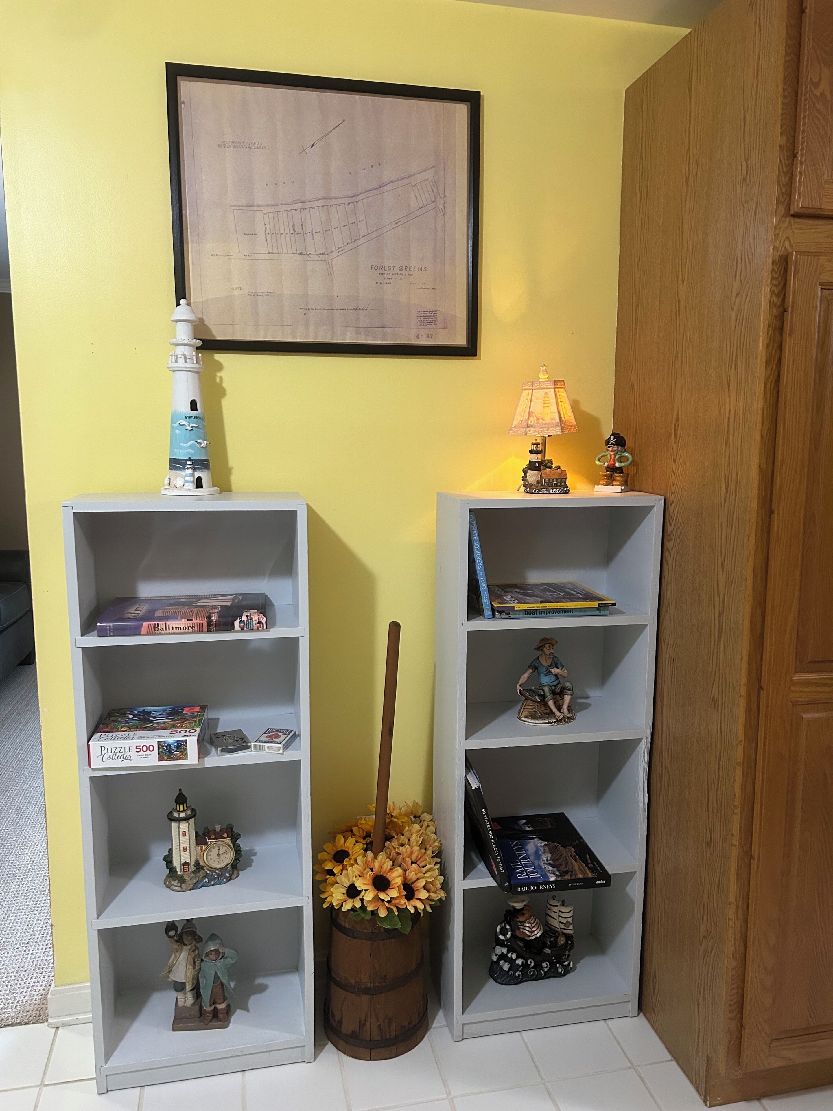 Une pièce comprenant deux étagères blanches, une carte encadrée, une maquette de phare, une lampe et un tonneau décoré de tournesols.