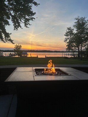 Un foyer avec des flammes, entouré d’une structure métallique, donnant sur une étendue d’eau avec une jetée et des arbres en arrière-plan.