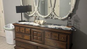A wooden dresser with a large ornate mirror above it, a lamp on the dresser, and a white laundry basket to the left.