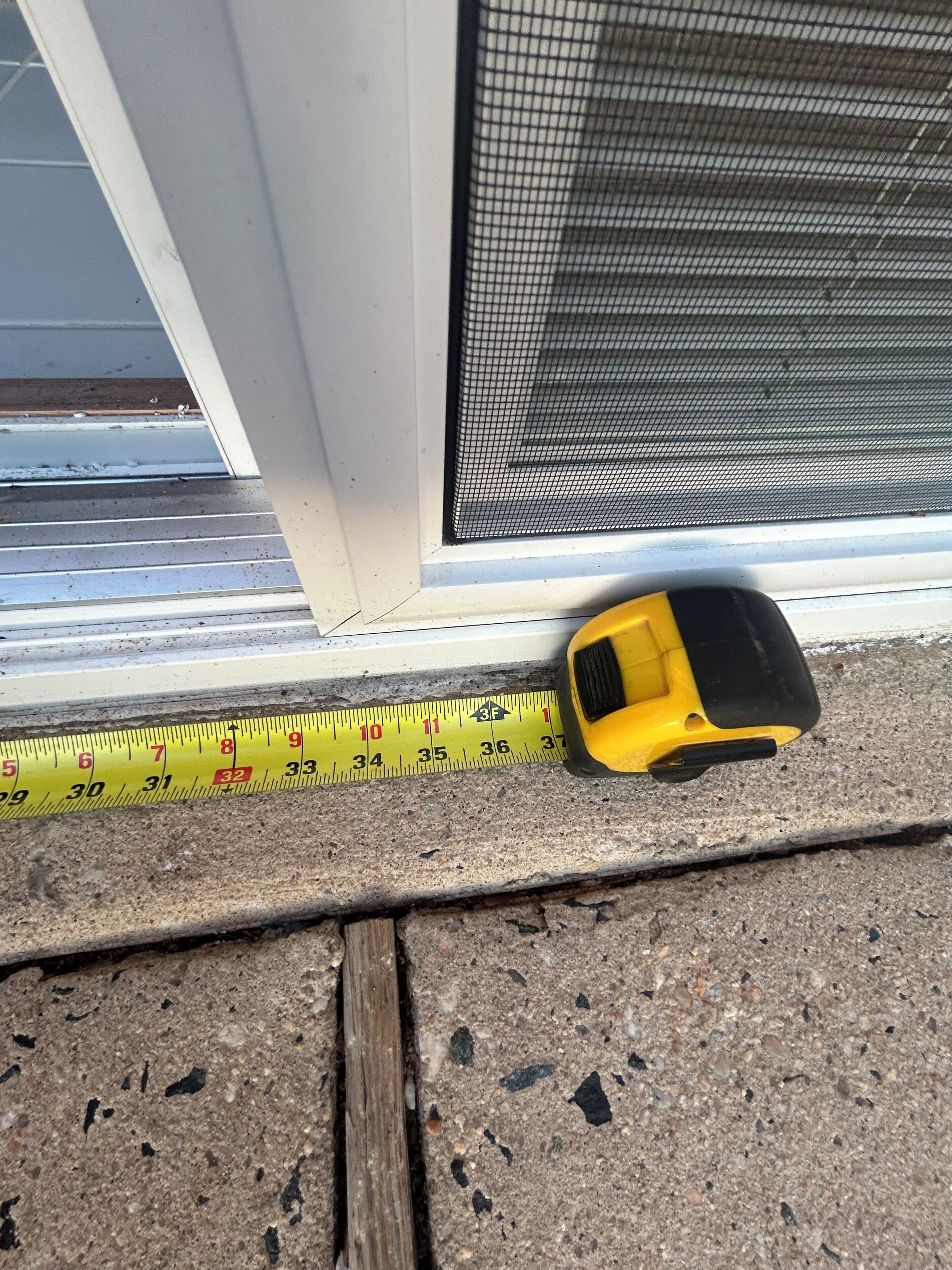 Un mètre ruban jaune est posé sur un sol en béton, à côté d’une fenêtre munie d’un grillage métallique.