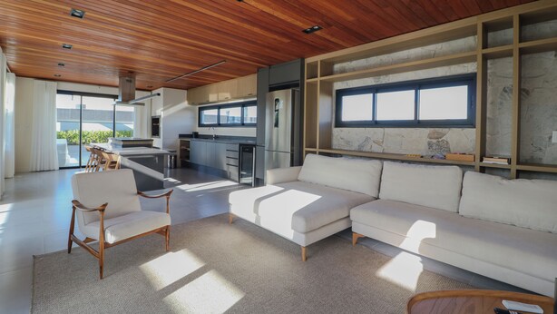 A modern living room with a wooden ceiling, a white sofa, a wooden armchair, and a kitchen in the background. - Complete Loft - New Campeche. Swimming pool, gym, laundry facilities, and close to the beach. (Florianópolis)
