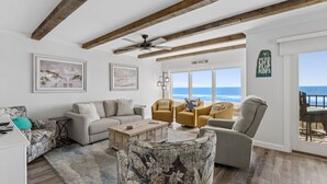 A living room with a view of the ocean, wooden beams, a ceiling fan, and a sign that says 'Live your life on the flip flops.'