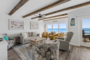 A living room with a view of the ocean, wooden beams, a ceiling fan, and a sign that says 'Live your life on the flip flops.'