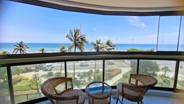 Un balcon avec des fauteuils en osier et une table, donnant sur une plage bordée de palmiers et l’océan.