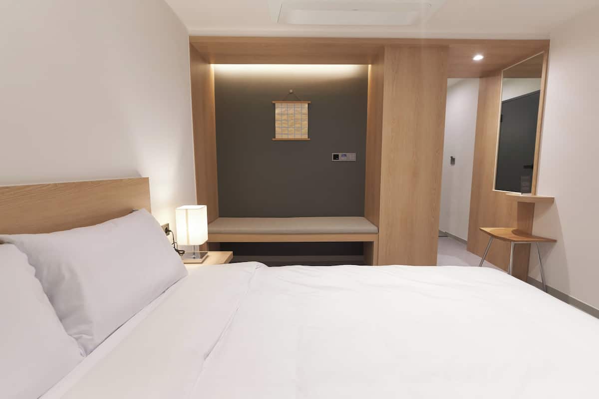 A hotel room with a bed, bedside lamp, a small bench, a mirror, and a wall-mounted light fixture.