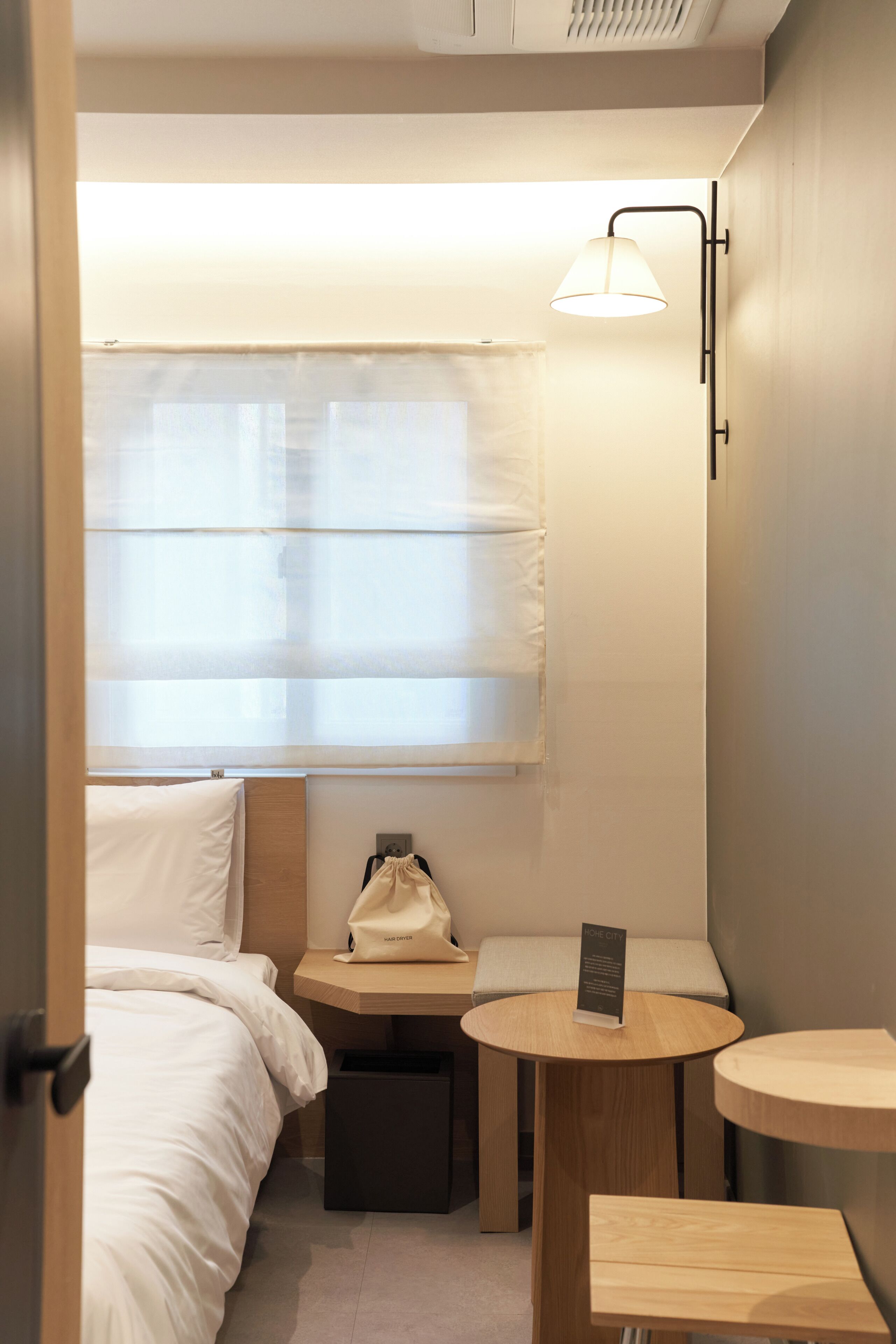 Ein kompaktes Hotelzimmer mit Bett, einer kleinen Sitzecke mit Tisch, einer Wandlampe und einem Fenster mit Jalousien.