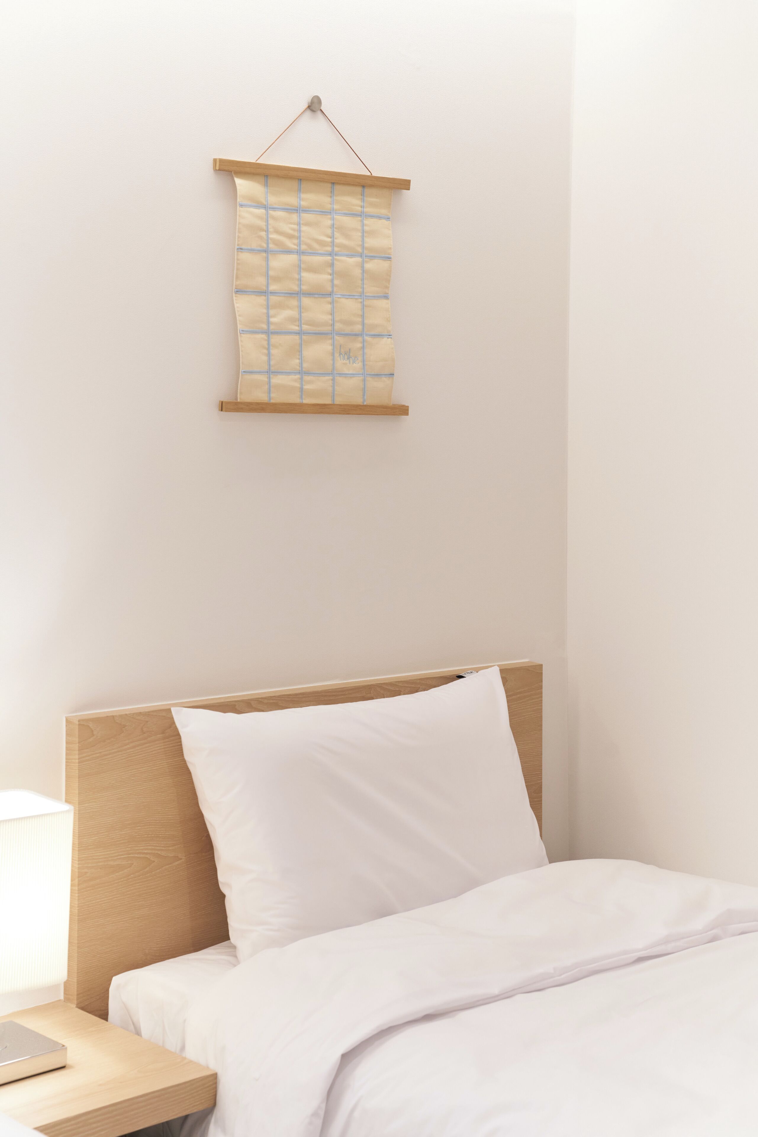 A neatly made bed with a wooden headboard, a bedside table with a lamp, and a wall-mounted hanging with a grid pattern.