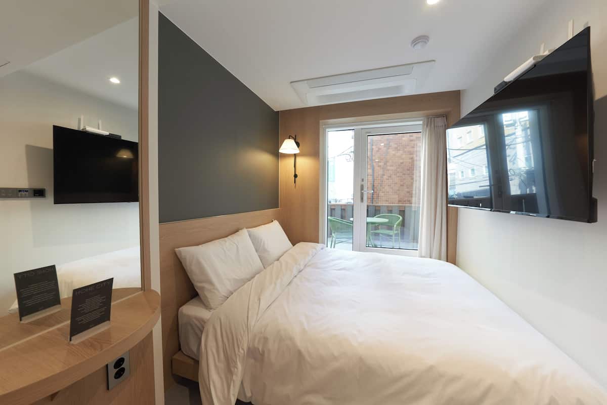 A hotel room with a bed, a TV, a window with a view of the outside, and a small table with a sign.