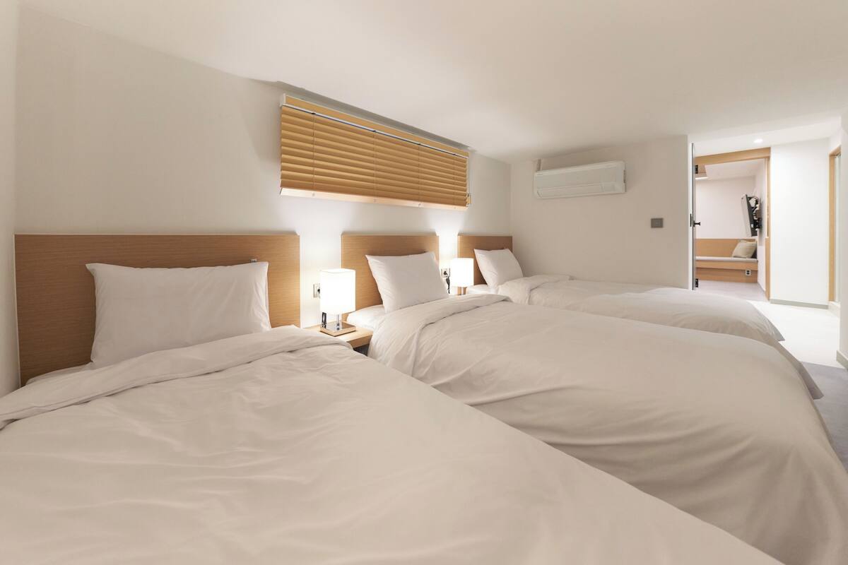 A hotel room with three beds, a wooden headboard, a window with blinds, a bedside table with a lamp, and a door leading to a bathroom.